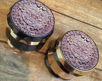May include: Two gold and black metal grinders with a detailed Aztec calendar design on the top. The grinders are made of metal and have a textured finish.