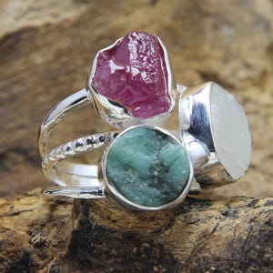 May include: A silver ring with three raw gemstones: a pink ruby, a green emerald, and a white moonstone.