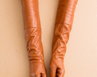 Op de afbeelding: Een paar lange operahandschoenen van bruin leer die tot voorbij de elleboog reiken. De handschoenen hebben een gladde, matte afwerking met zichtbare stiksels langs de vingers en een horizontale naad net boven het polsgedeelte van elke handschoen.