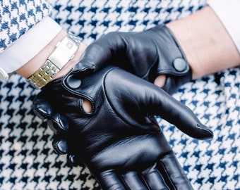 May include: A pair of black leather gloves featuring a small circular cutout and a snap button closure at the wrist. The gloves are worn with a silver-toned metal link watch and a white and navy blue houndstooth patterned garment.