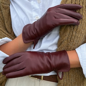 Könnte beinhalten: Ein Paar lange, burgunderfarbene Lederhandschuhe mit einem Umschlag. Die Handschuhe sind aus weichem, geschmeidigem Leder gefertigt und haben einen klassischen, eleganten Look.