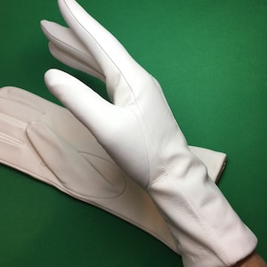 Könnte beinhalten: Ein Paar weiße Lederhandschuhe. Die Handschuhe sind aus glattem, geschmeidigem Leder gefertigt und haben ein klassisches Design. Die Handschuhe sind auf grünem Hintergrund abgebildet.