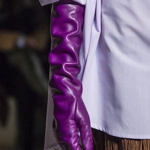 Könnte beinhalten: Nahaufnahme eines Arms mit einem langen, leuchtend violetten Lederhandschuh. Der Handschuh ist zerknittert und strukturiert. Die Person trägt ein weißes Hemd mit hohem Kragen und einen braunen Fransenrock.