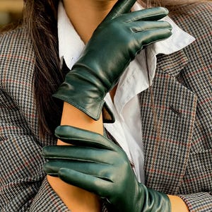 Könnte beinhalten: Ein Paar dunkelgrüne Lederhandschuhe. Die Handschuhe werden mit einem weißen Hemd und einem grau-braunen Blazer mit Karomuster getragen. Die Handschuhe sind einfarbig und scheinen aus weichem Leder zu sein.