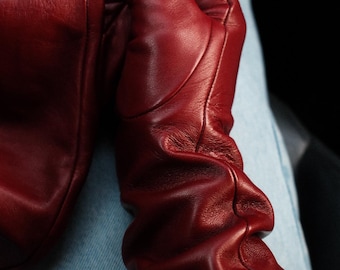 May include: A pair of deep red leather gloves featuring a smooth finish and a gathered, ruched design along the wrist area. The gloves are constructed with visible stitched seams and have a soft, flexible texture that contours to the hand.