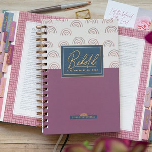 bible-journal-for-verse-by-verse-bible-study-with-rainbows-on-hard-cover-laying-on-open-bible-with-ombre-bible-tabs-and-cursive-gold-lettering