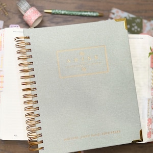 May include: A sage green spiral-bound planner with gold wire binding and gold corner protectors. The cover features the word ARISE in gold foil lettering within a rectangular border, with additional small gold text printed at the bottom edge.
