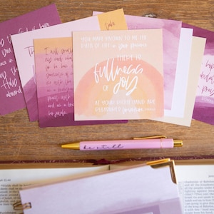 May include: A set of pink and orange watercolor cards with inspirational quotes and bible verses. The cards feature a variety of designs, including a rainbow, a sunset, and a floral pattern. The text on the cards includes quotes such as "You make known to me the path of life; in your presence there is fullness of joy" and "Hope, be steadfast; prayer, be constant."