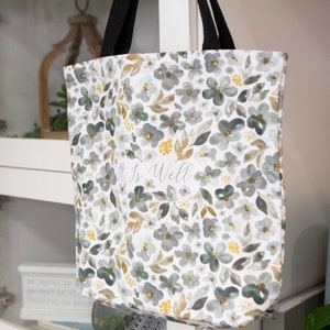 May include: A white canvas tote bag with black handles and a floral pattern in shades of gray, brown, and yellow. The bag has the text "It is Well" printed on the front.