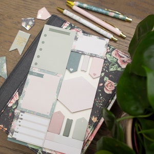 May include: A set of floral patterned sticky notes and notepads in pink and green colors. The notes are arranged on a black floral patterned notebook.