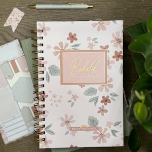 May include: A pink and white floral patterned journal with a gold spiral binding. The cover has a pink rectangle with the text "Behold I am with you always" in gold lettering. The bottom of the cover says "Bible Study Journal".