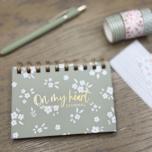 May include: A small, spiral-bound journal with a floral pattern and the words "On my heart" in gold script. A matching pen, decorative tape, and patterned cards are also present, all in coordinating colors.