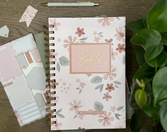 May include: A pink and white floral patterned journal with a gold spiral binding. The cover has a pink rectangle with the text "Behold I am with you always" in gold lettering. The bottom of the cover says "Bible Study Journal".