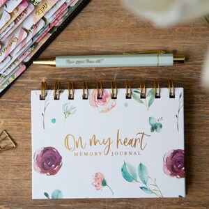 May include: A white spiral bound journal with a floral design and gold lettering that reads "On my heart MEMORY JOURNAL". The journal is on a wooden surface with a pen, tape, and paper clips.