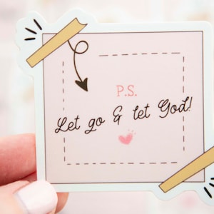 May include: A pink and white sticker with a handwritten message that says "P.S. Let go & let God!" The sticker has a brown tape design on the top and bottom and a small pink heart in the center.
