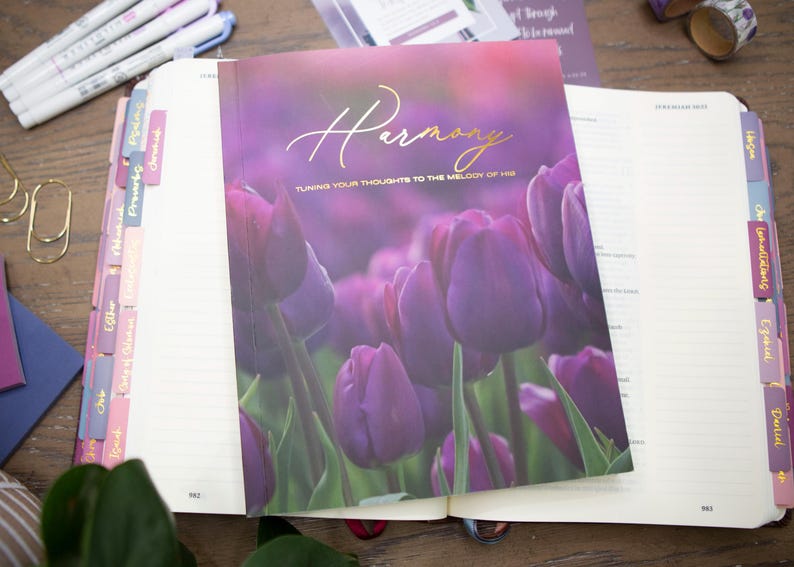 May include: A journal titled "Harmony" with a purple floral cover featuring tulips. The journal is open, revealing lined pages and colorful tabs. Pens, paperclips, and washi tape are visible on the wooden surface.