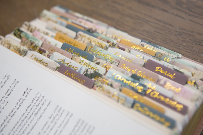 Puede incluir: Primer plano de una Biblia con pesta&ntilde;as florales de colores que marcan cada libro de la Biblia. Las pesta&ntilde;as est&aacute;n etiquetadas con los nombres de los libros, incluyendo &Eacute;xodo, Jueces, 2 Reyes, Ezequiel, Cantar de los Cantares, Daniel, 1 Cr&oacute;nicas y 2 Cr&oacute;nicas.