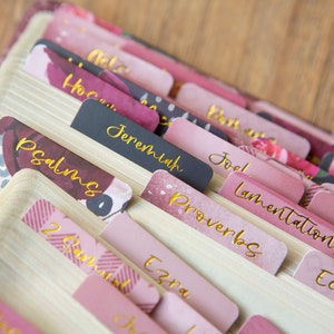May include: Pink and black book tabs with gold lettering for the books of the Bible, including Psalms, Jeremiah, Proverbs, Lamentations, Joel, Ecclesiastes, Ezra, and Amos.