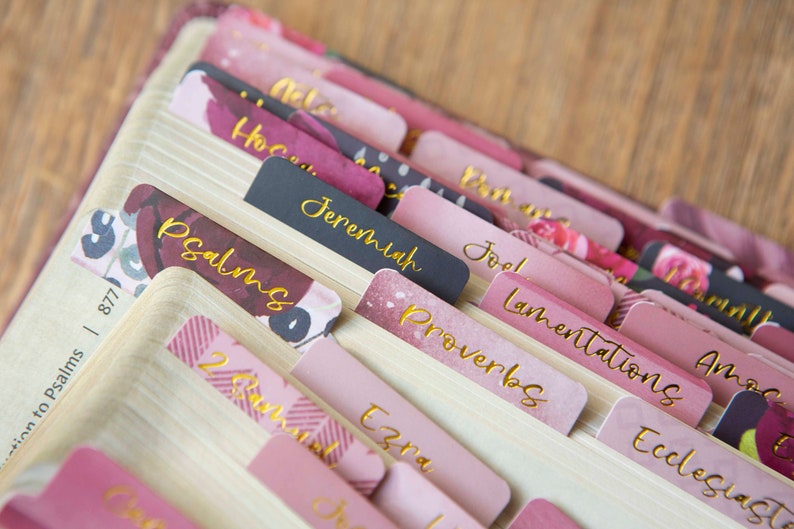 May include: Pink and black book tabs with gold lettering for the books of the Bible, including Psalms, Jeremiah, Proverbs, Lamentations, Joel, Ecclesiastes, Ezra, and Amos.