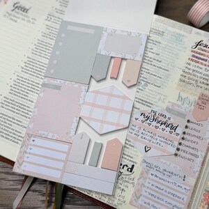 May include: A set of adhesive sticky notes in shades of soft pink, grey, and white, featuring various shapes including rectangles, hexagons, and arrow-shaped tabs, placed on an open book with handwritten notes and decorative washi tape.