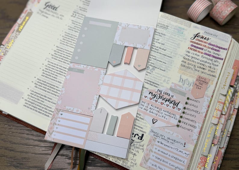 May include: A set of adhesive sticky notes in shades of soft pink, grey, and white, featuring various shapes including rectangles, hexagons, and arrow-shaped tabs, placed on an open book with handwritten notes and decorative washi tape.