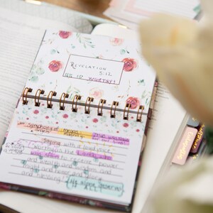 May include: A floral patterned journal with a gold spiral binding. The journal has a white background with pink and green watercolor flowers. The text "Revelation 5:12 He is worthy!" is written in black marker on the front cover.