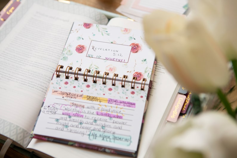 May include: A floral patterned journal with a gold spiral binding. The journal has a white background with pink and green watercolor flowers. The text "Revelation 5:12 He is worthy!" is written in black marker on the front cover.