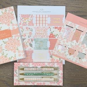 May include: A stationery set featuring a floral notebook labeled Selah Sticky Notes, a sheet of peach and green patterned adhesive tabs, a set of eight magnetic bookmarks with floral and solid designs, and three pens in a floral box by Chosen & Cherished.