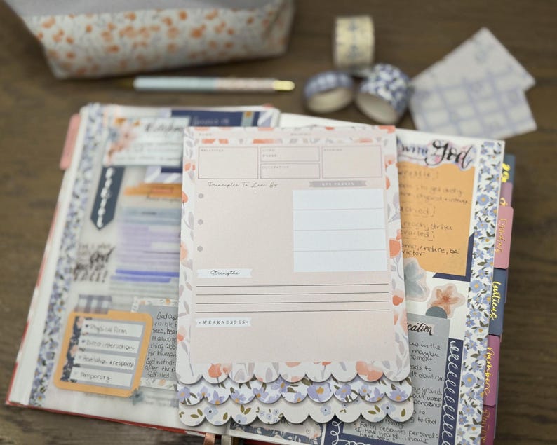 May include: A planner with a pink and floral design, open to a page with a pink paper insert. The insert has a scalloped edge and the text "Principles to Live By". Decorative tape, a pen, and a pencil case are in the background.