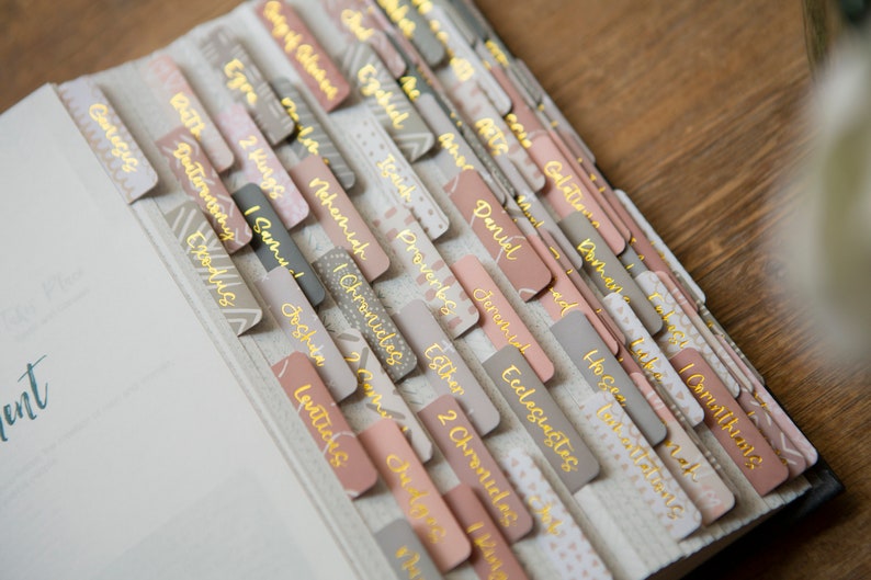 May include: A close-up of a Bible with pink and gray tabs labeled with the names of books of the Bible, including Genesis, Exodus, Leviticus, Numbers, Deuteronomy, Joshua, Judges, Ruth, 1 Samuel, 2 Samuel, 1 Kings, 2 Kings, 1 Chronicles, 2 Chronicles, Ezra, Nehemiah, Esther, Job, Psalms, Proverbs, Ecclesiastes, Song of Solomon, Isaiah, Jeremiah, Lamentations, Ezekiel, Daniel, Hosea, Joel, Amos, Obadiah, Jonah, Micah, Nahum, Habakkuk, Zephaniah, Haggai, Zechariah, Malachi, Matthew, Mark, Luke, John, Acts, Romans, 1 Corinthians, 2 Corinthians, Galatians, Ephesians, Philippians, Colossians, 1 Thessalonians, 2 Thessalonians, 1 Timothy, 2 Timothy, Titus, Philemon, Hebrews, James, 1 Peter, 2 Peter, 1 John, 2 John, 3 John, Jude, and Revelation.