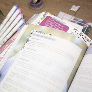 May include: An open book with text and floral accents, alongside a stack of colorful pens and a purple journal. The book's pages are filled with text, and a bookmark reads "save the Lord with her".