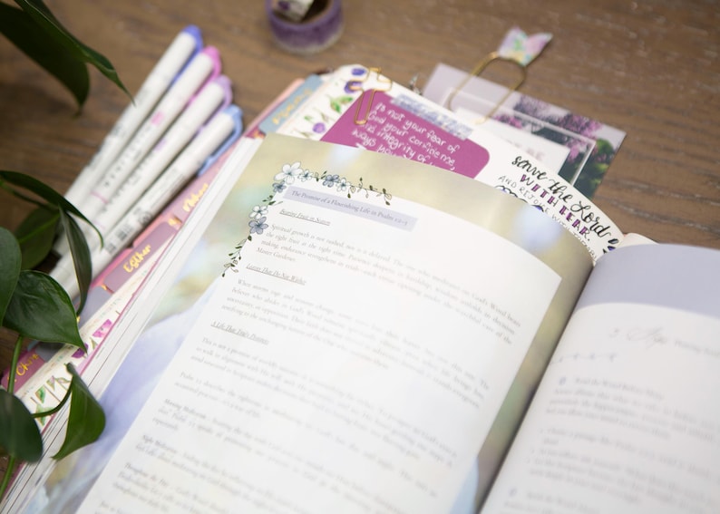 May include: An open book with text and floral accents, alongside a stack of colorful pens and a purple journal. The book's pages are filled with text, and a bookmark reads "save the Lord with her".