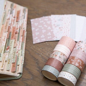 Pode incluir: Um livro aberto com abas rotuladas e rolos de fita adesiva decorativa. Os rolos de fita adesiva apresentam desenhos florais e estampados em tons de rosa, castanho e verde. Uma pilha de papel estampado tamb&eacute;m &eacute; vis&iacute;vel.