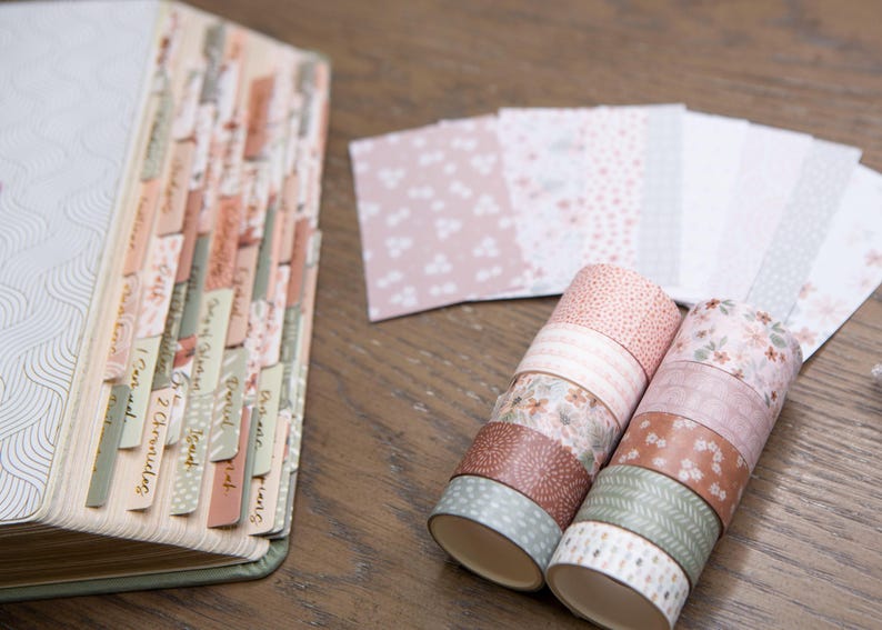 Pode incluir: Um livro aberto com abas rotuladas e rolos de fita adesiva decorativa. Os rolos de fita adesiva apresentam desenhos florais e estampados em tons de rosa, castanho e verde. Uma pilha de papel estampado tamb&eacute;m &eacute; vis&iacute;vel.