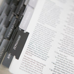 May include: A close-up of a Bible with black and gray tabs marking different books of the Bible. The tabs are labeled "Job", "Isaiah", "Jon", "Zephaniah", "Song of Solomon", "Esther", "Ecclesiastes", and "Habakkuk".