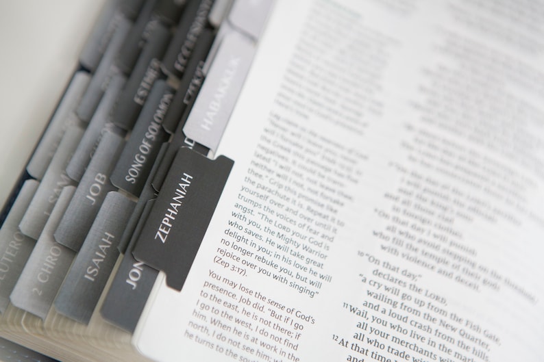 May include: A close-up of a Bible with black and gray tabs marking different books of the Bible. The tabs are labeled "Job", "Isaiah", "Jon", "Zephaniah", "Song of Solomon", "Esther", "Ecclesiastes", and "Habakkuk".