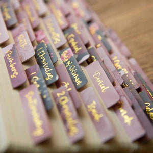 May include: A close-up of a stack of pink and black book dividers with gold lettering. The dividers are labeled with names of books from the Bible, including Numbers, 1 Chronicles, Song of Solomon, Daniel, Isaiah, and Colossians.