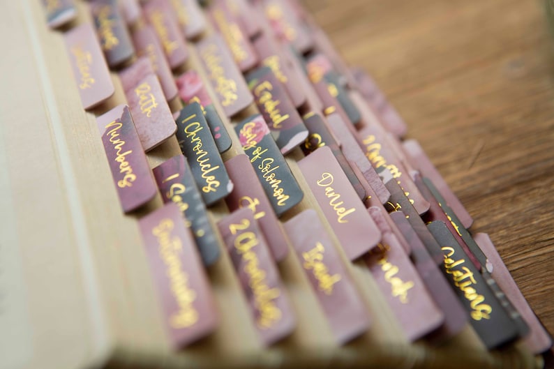 May include: A close-up of a stack of pink and black book dividers with gold lettering. The dividers are labeled with names of books from the Bible, including Numbers, 1 Chronicles, Song of Solomon, Daniel, Isaiah, and Colossians.