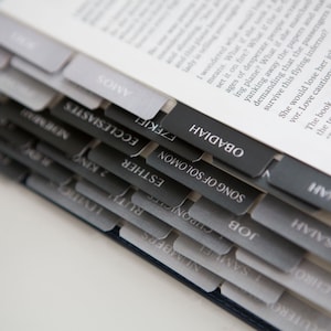 May include: A close-up of a book with black and gray tabs labeled with the names of books of the Bible, including "Song of Solomon", "Job", "Isaiah", "Jonah", "Obadiah", "Amos", "Ecclesiastes", "Ezekiel", and "Esther".