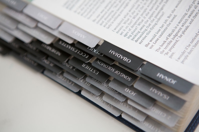 May include: A close-up of a book with black and gray tabs labeled with the names of books of the Bible, including "Song of Solomon", "Job", "Isaiah", "Jonah", "Obadiah", "Amos", "Ecclesiastes", "Ezekiel", and "Esther".