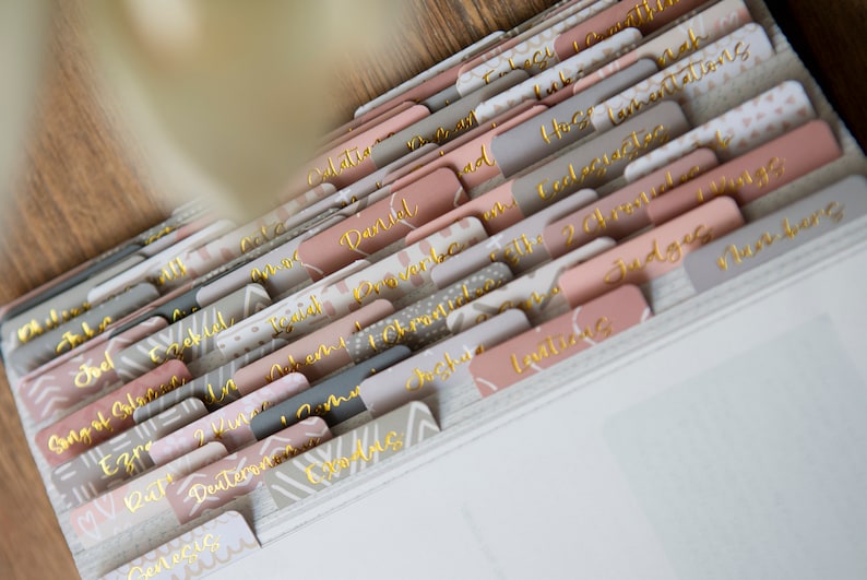 May include: A set of colorful, patterned dividers with gold lettering for a binder. The dividers are labeled with different books of the Bible, including Genesis, Exodus, Leviticus, Numbers, Deuteronomy, Joshua, Judges, Ruth, 1 Samuel, 2 Samuel, 1 Kings, 2 Kings, 1 Chronicles, 2 Chronicles, Ezra, Nehemiah, Esther, Job, Psalms, Proverbs, Ecclesiastes, Song of Solomon, Isaiah, Jeremiah, Lamentations, Ezekiel, Daniel, Hosea, Joel, Amos, Obadiah, Jonah, Micah, Nahum, Habakkuk, Zephaniah, Haggai, Zechariah, Malachi, Matthew, Mark, Luke, John, Acts, Romans, 1 Corinthians, 2 Corinthians, Galatians, Ephesians, Philippians, Colossians, 1 Thessalonians, 2 Thessalonians, 1 Timothy, 2 Timothy, Titus, Philemon, Hebrews, James, 1 Peter, 2 Peter, 1 John, 2 John, 3 John, Jude, and Revelation.