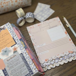 May include: A planner and stationery set with a floral theme. Includes a notebook, decorative tape rolls, notepads, and a pen. The notebook has handwritten notes and floral accents. The notepads have a scalloped edge and floral design.