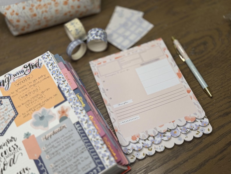 May include: A planner and stationery set with a floral theme. Includes a notebook, decorative tape rolls, notepads, and a pen. The notebook has handwritten notes and floral accents. The notepads have a scalloped edge and floral design.