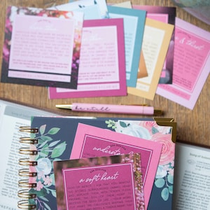 May include: A close-up of a pink card with the text "Ask God for a soft heart" and a floral background. The card is on top of a blue and floral patterned journal with a gold spiral binding. A pink pen is lying on top of the journal.