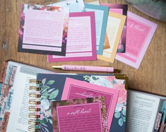 May include: A close-up of a pink card with the text "Ask God for a soft heart" and a floral background. The card is on top of a blue and floral patterned journal with a gold spiral binding. A pink pen is lying on top of the journal.