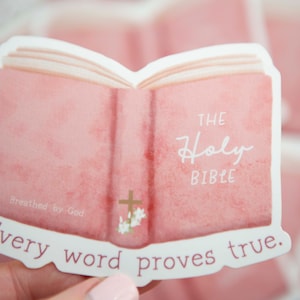 May include: A pink watercolor sticker of an open book with the words "The Holy Bible" and "Every word proves true." The sticker has a gold cross and flowers on the front cover.