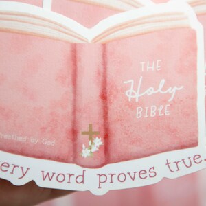May include: A pink sticker with an open book illustration. The book has the words "The Holy Bible" written on it. The sticker also has the text "Breathed by God" and "Every word proves true."