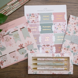Peut inclure: Un ensemble de papeterie &agrave; th&egrave;me floral rose et vert comprenant un carnet, des stylos, des signets et des autocollants. Le carnet porte le texte "Selah" et "Scripture Notes" sur la couverture. Les autocollants ont diverses phrases dont "Tourne tes yeux vers J&eacute;sus" et "Je peux tout faire par le Christ."