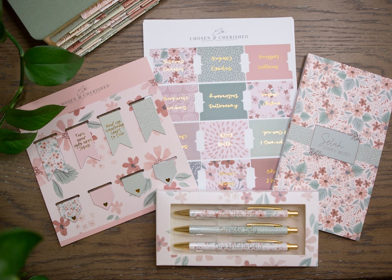 Peut inclure: Un ensemble de papeterie &agrave; th&egrave;me floral rose et vert comprenant un carnet, des stylos, des signets et des autocollants. Le carnet porte le texte "Selah" et "Scripture Notes" sur la couverture. Les autocollants ont diverses phrases dont "Tourne tes yeux vers J&eacute;sus" et "Je peux tout faire par le Christ."