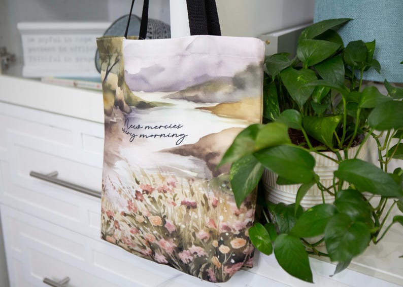 Puede incluir: Una bolsa de tela blanca con un dise&ntilde;o de paisaje de acuarela. La bolsa tiene un asa negra y el texto "New mercies every morning" impreso en la parte delantera.